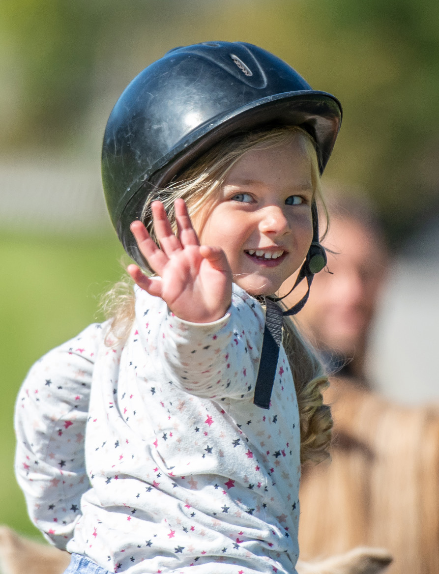 A young girl in a black riding helmet waving happily.