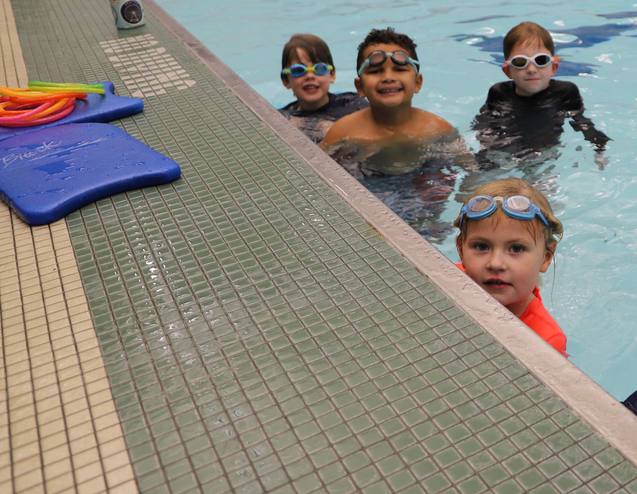 Four children with swimming goggles smiling at the edge of a pool