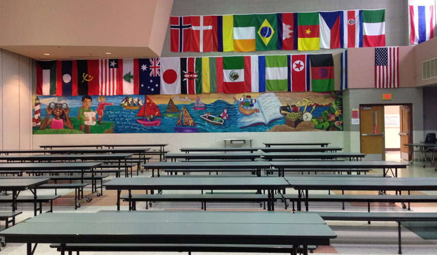 A large school cafeteria with rows of tables and international flags hanging from the ceiling
