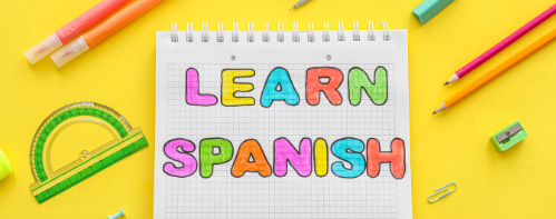 A young girl holding a sign that says 'Espa&ntilde;ol' next to a notebook that says 'LEARN SPANISH' surrounded by colorful pencils and a protractor.