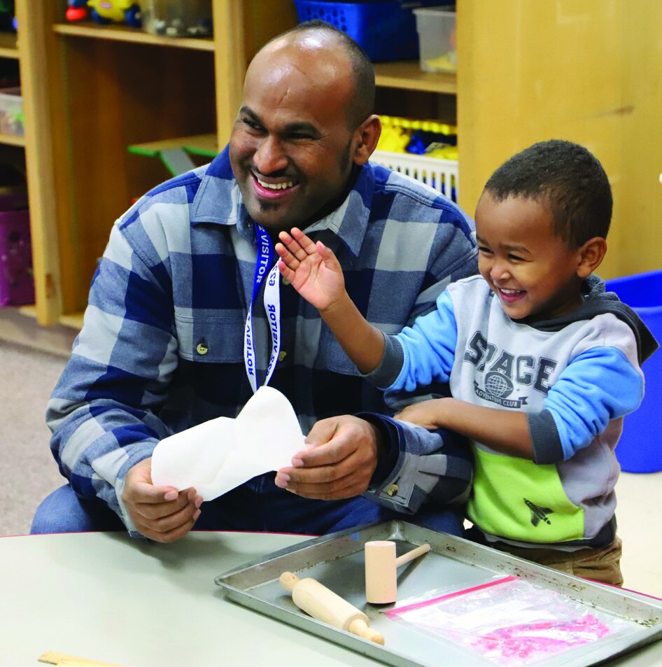 A smiling man and a young boy happily playing with dough and kitchen tools like a rolling pin and wooden mallet at a table.