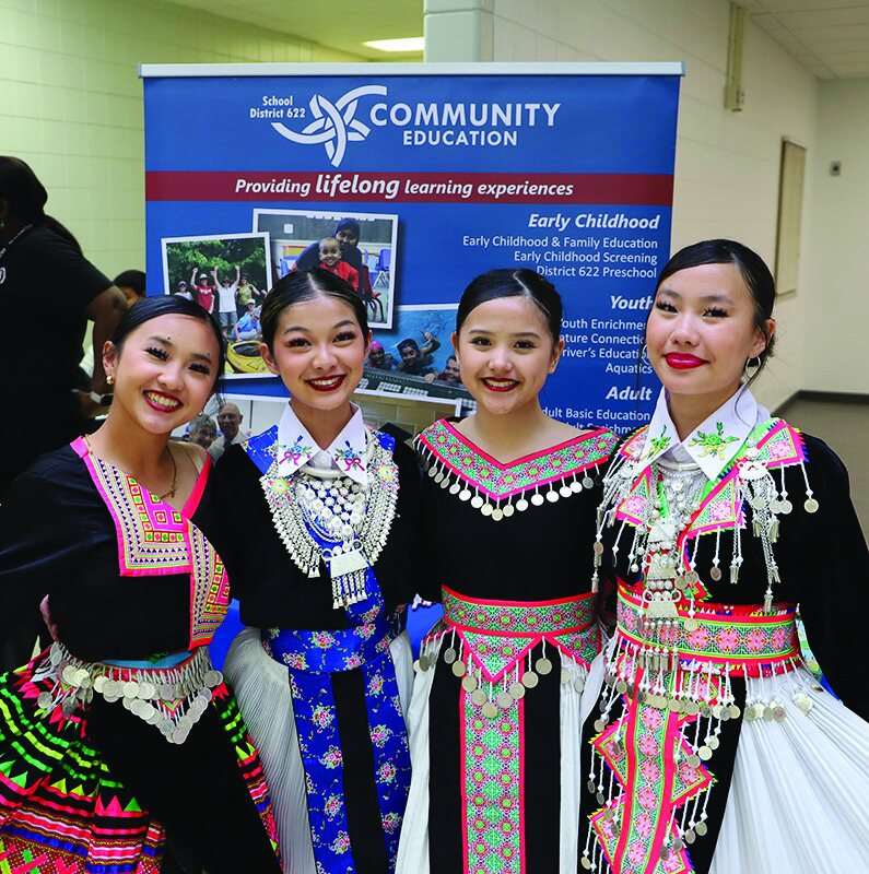 Four young women in vibrant traditional Hmong attire standing together in front of a banner for Community Education.