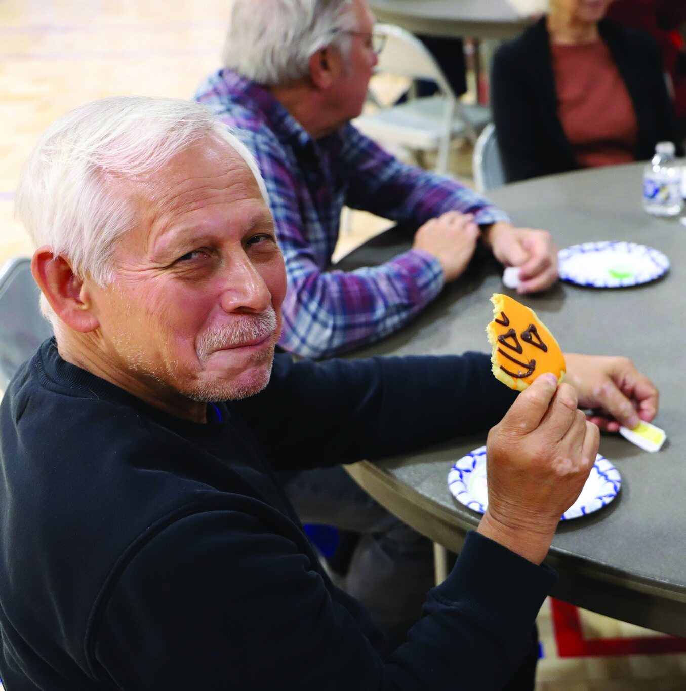 An elderly man sitting at a table, smiling while holding up a small orange pumpkin-shaped decoration.