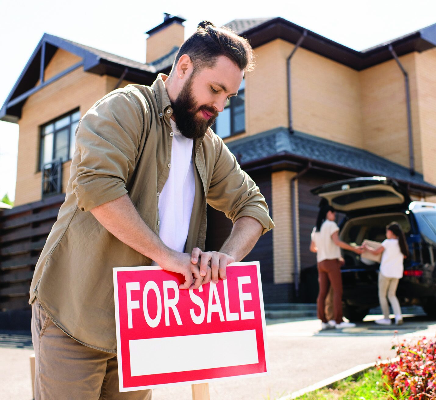 A man leaning over a red and white 'FOR SALE' sign in front of a modern house, while a family packs a car in the background.