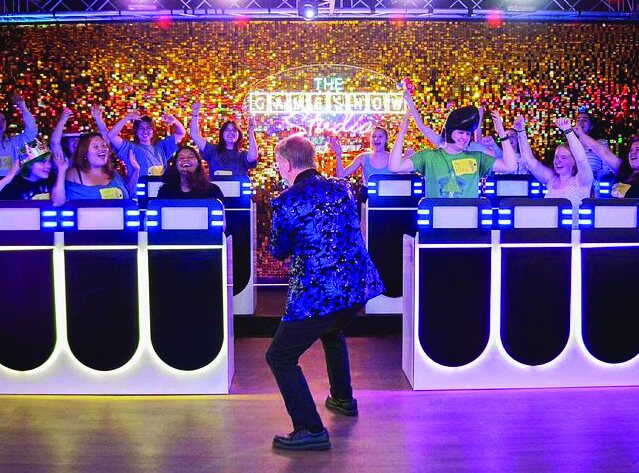 A vibrant group photo from 'The Gameshow Studio' where a host in a blue jacket stands before contestants at themed podiums on a brightly lit stage.