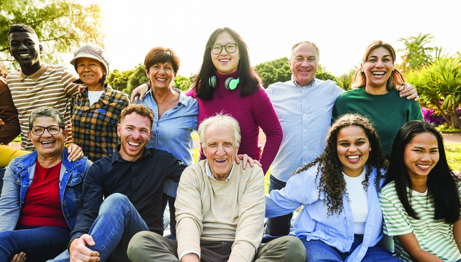 A large, diverse group of smiling people of various ages and ethnicities posing together outdoors on a sunny day.