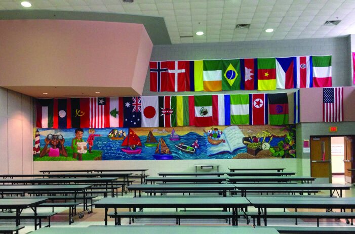 A school cafeteria with long tables and colorful international flags hanging from the ceiling.