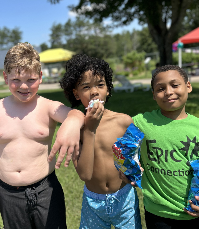 Three young boys posing outdoors, with one eating snacks and another wearing a green EPIC Connection shirt.