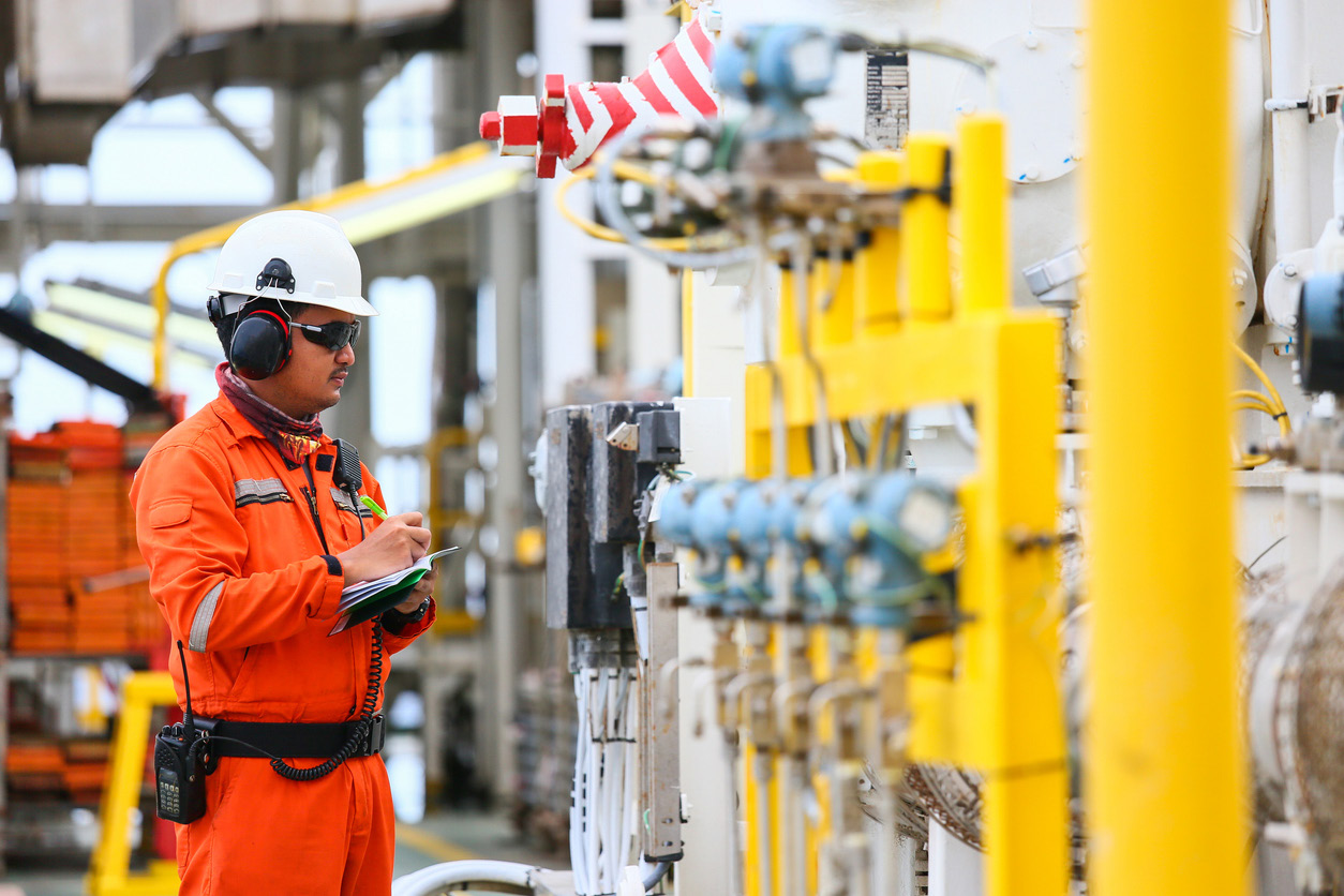 A worker in an orange jumpsuit writing on a notepad while standing next to industrial equipment
