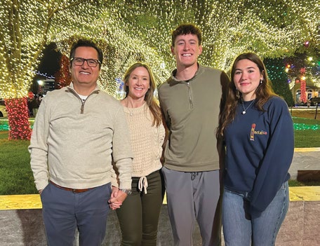 Family portrait with two adults and two young adults standing together in front of warm string lights