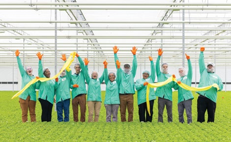 A group of employees in green uniforms standing inside a greenhouse, celebrating while holding a yellow ribbon