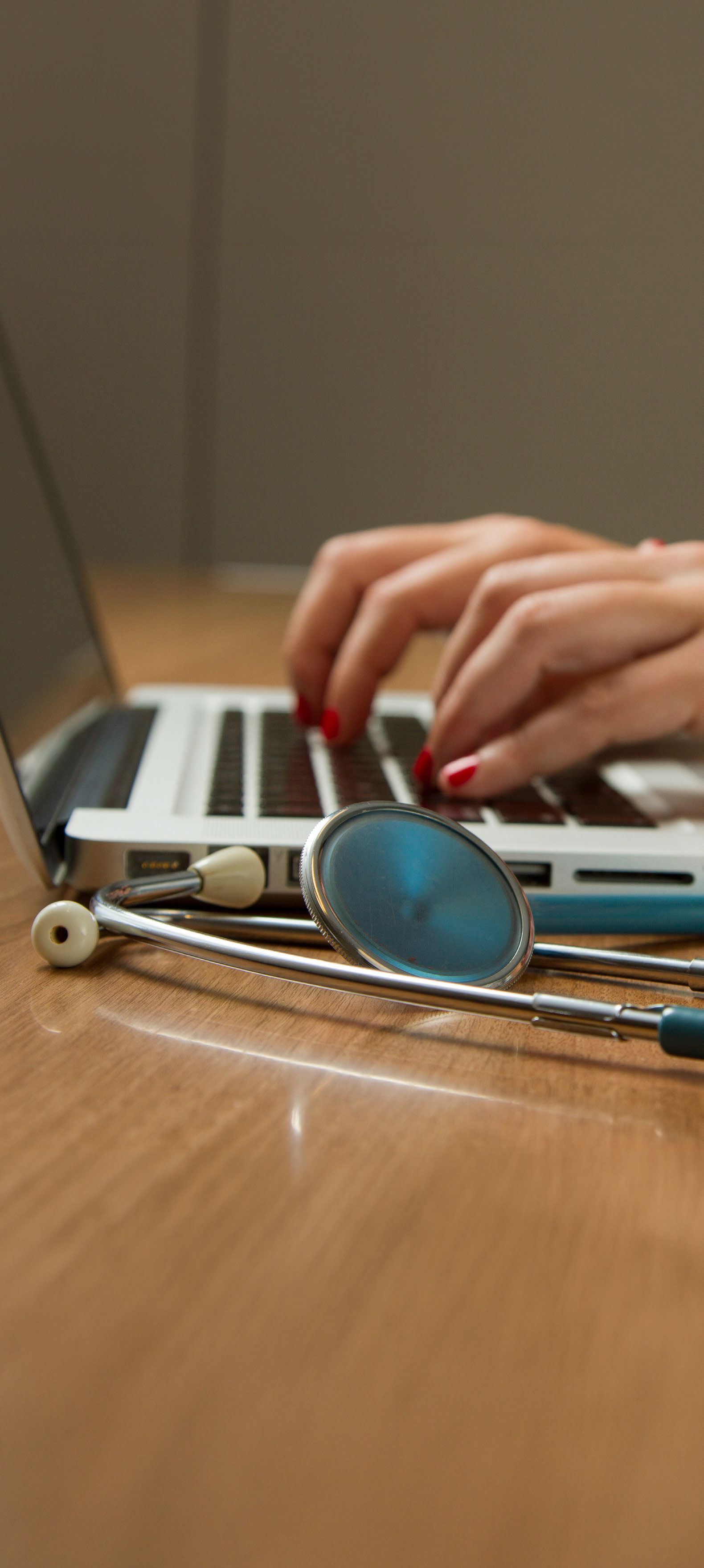 A close-up image of a person's hands with red-painted nails typing on a laptop keyboard with a stethoscope in the foreground.
