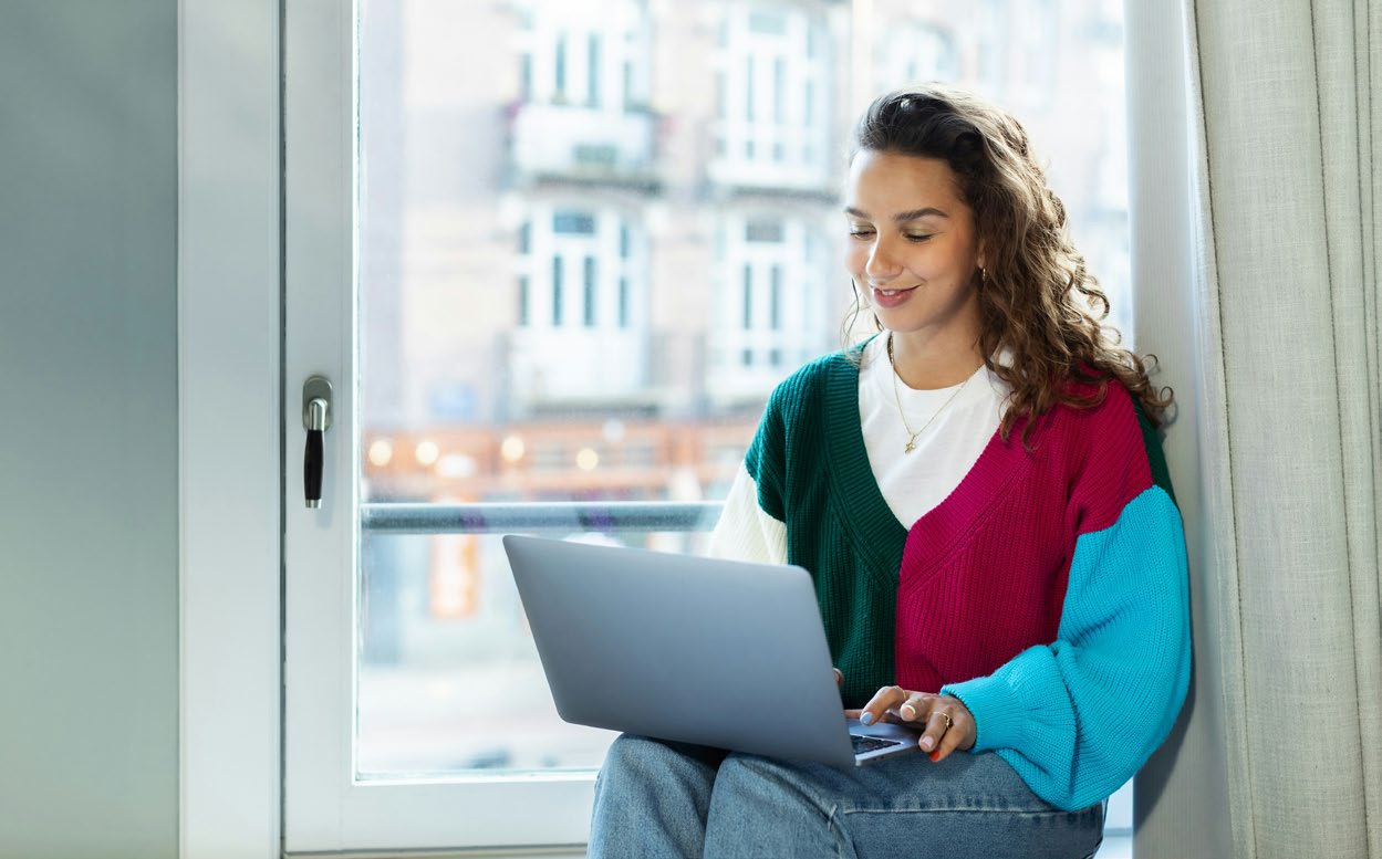 A young woman sitting on a windowsill, looking at her laptop screen.