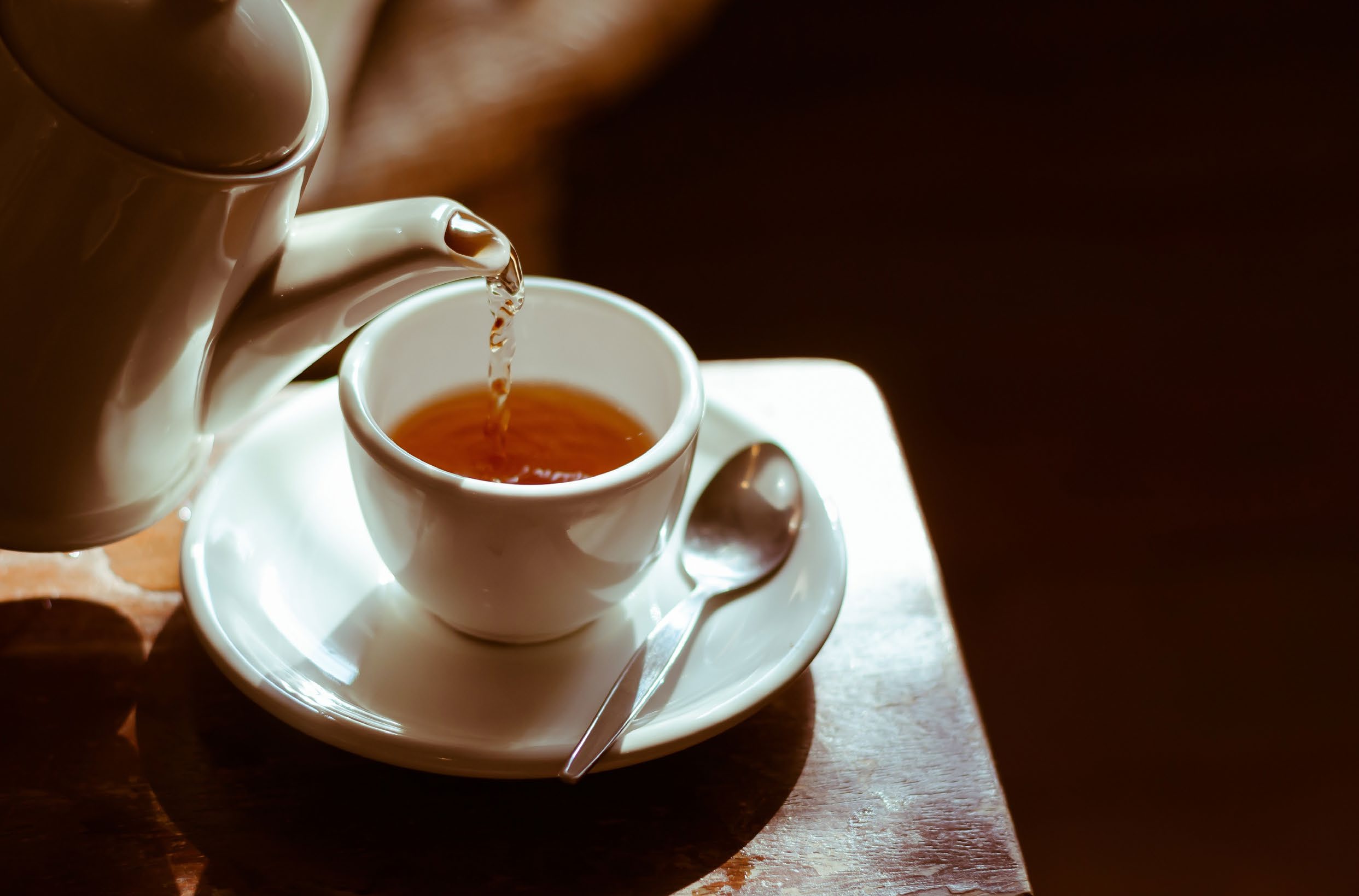 A high-angle shot of tea being poured from a white ceramic teapot into a white cup sitting on a saucer on a wooden table.