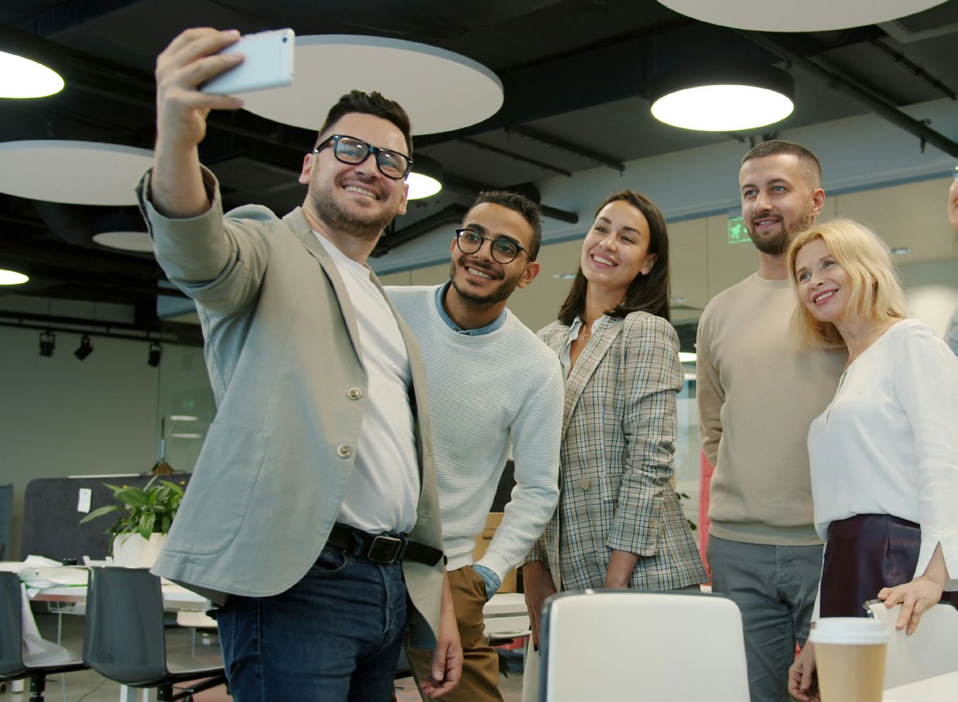 A group of diverse office colleagues gathered together, smiling and taking a selfie in their workplace.