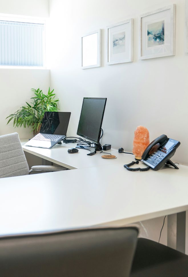 A clean and organized office workspace featuring a white desk with a monitor, plant, and lamp, against a wall with framed art.