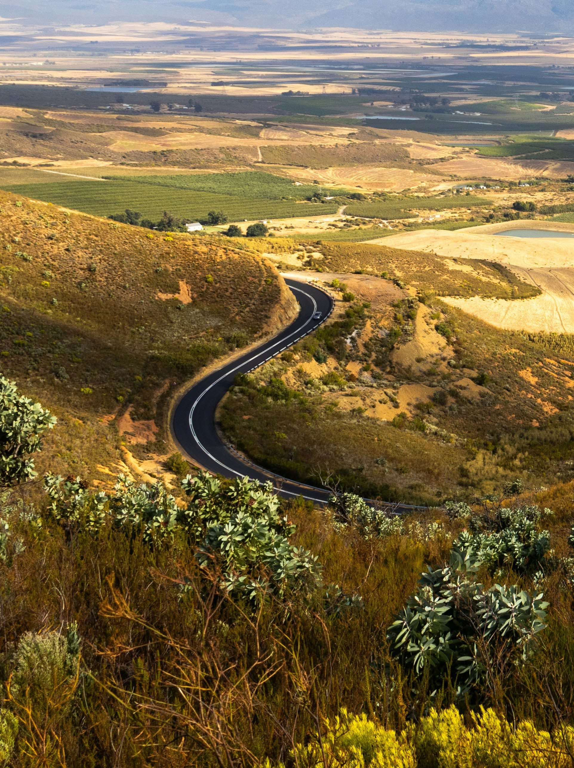 A high-angle landscape photograph showing a winding black asphalt road snaking through a vast, hilly valley.