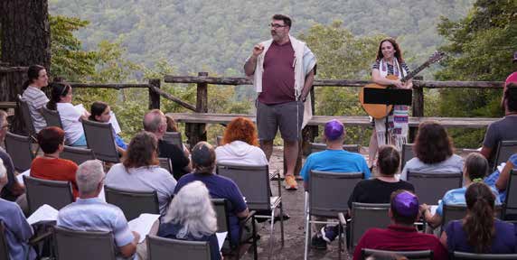 Photo of group of people gathered outdoors, speaking and listening, mountains and trees in the background.