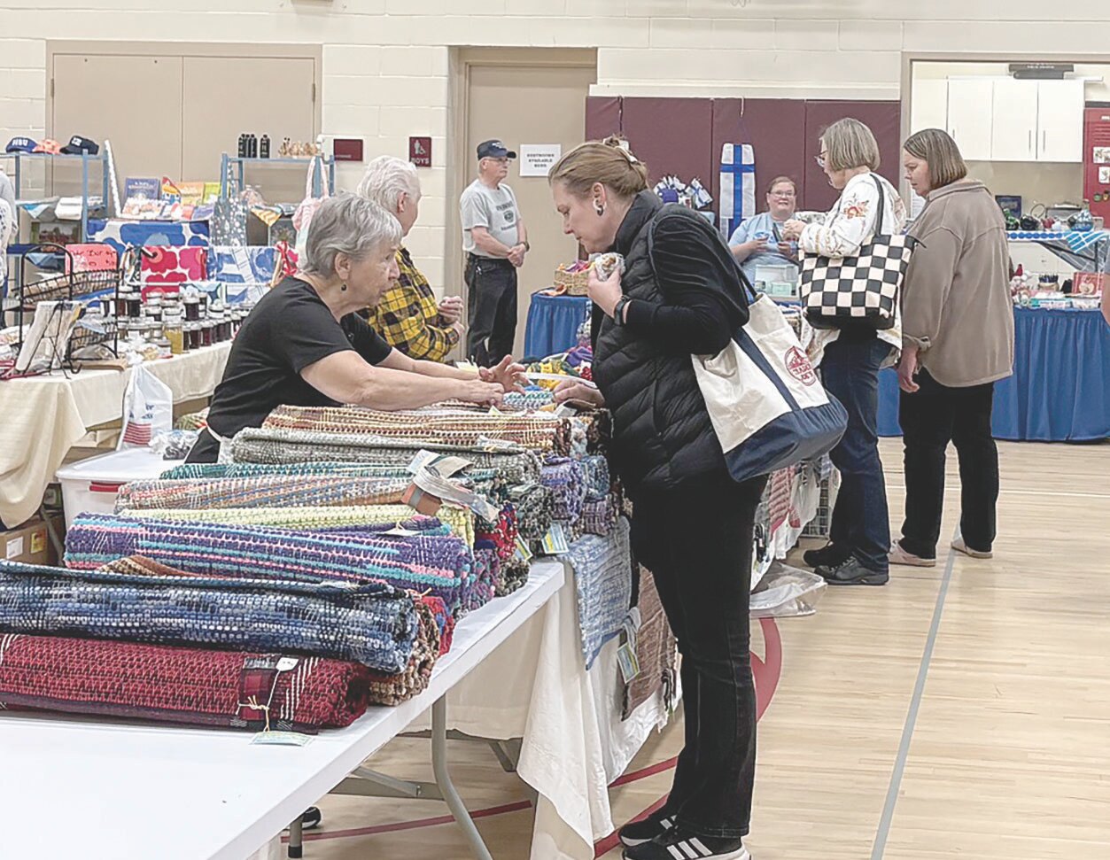 People browsing colorful, rolled-up hand-woven rugs displayed on a long table at an indoor market or tori during a festival.