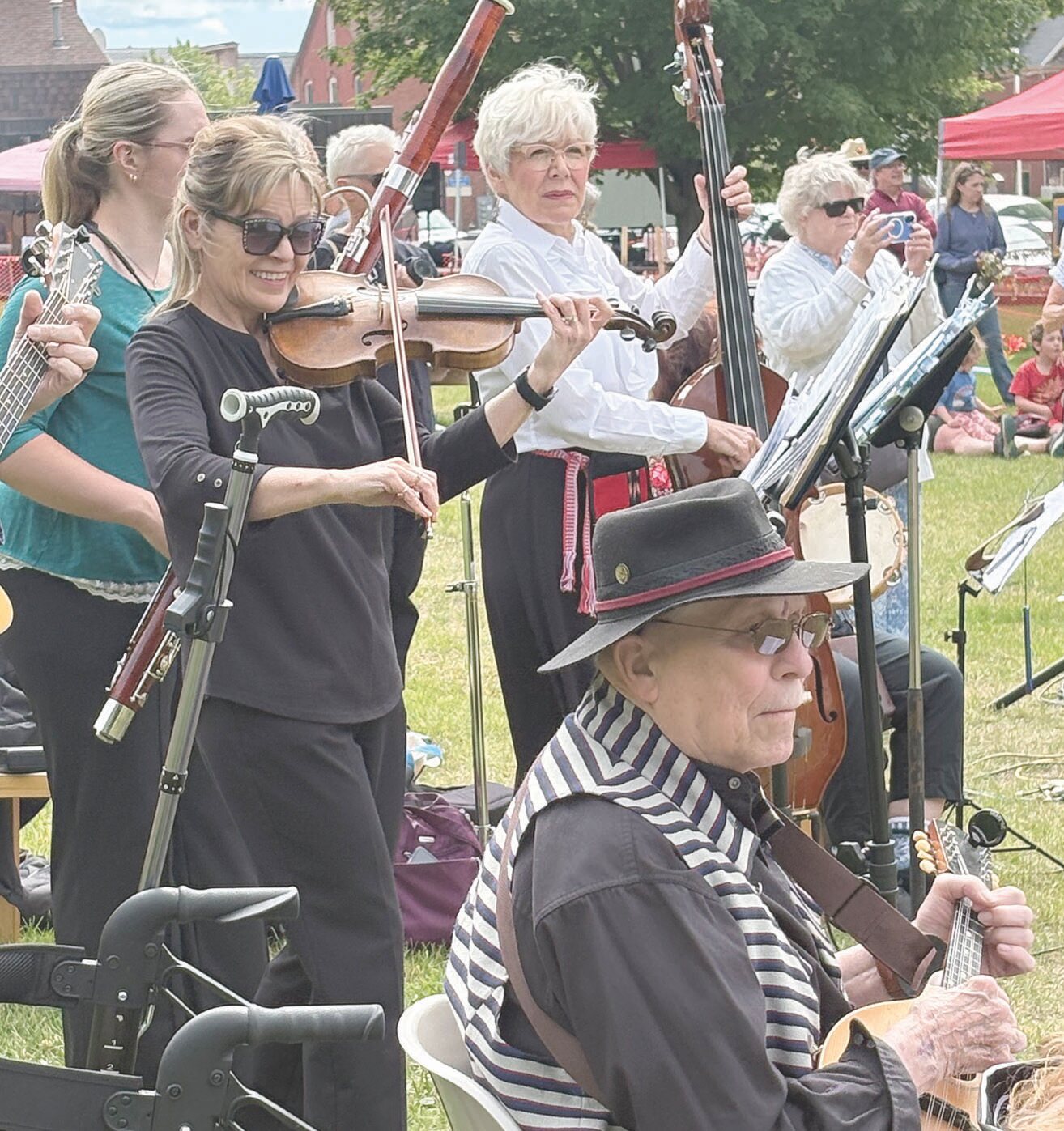 A group of musicians performing outdoors; in the foreground, a man in a hat and striped shirt plays an instrument, while in the background, several women play violins and a double bass.