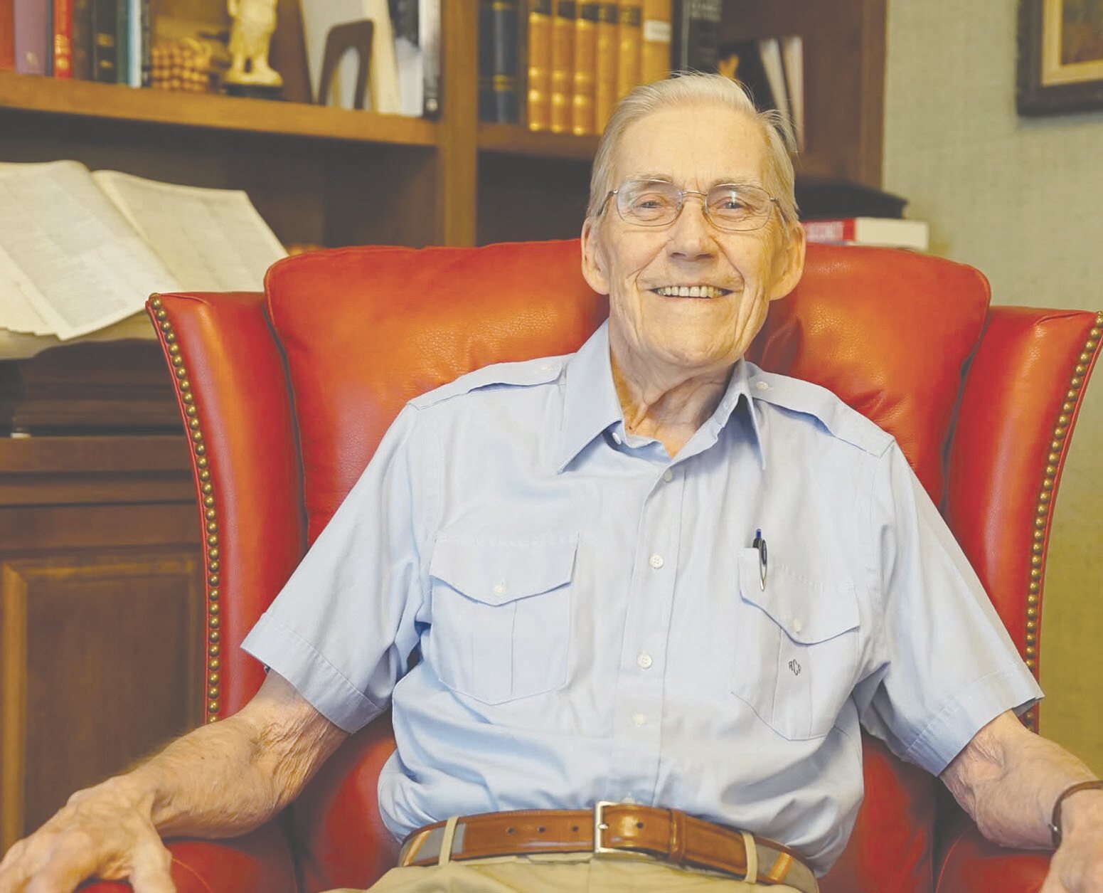 A portrait of Alpo Franssila Crane as an older man, wearing a light blue button-down shirt and glasses, smiling while sitting in a large red leather armchair.