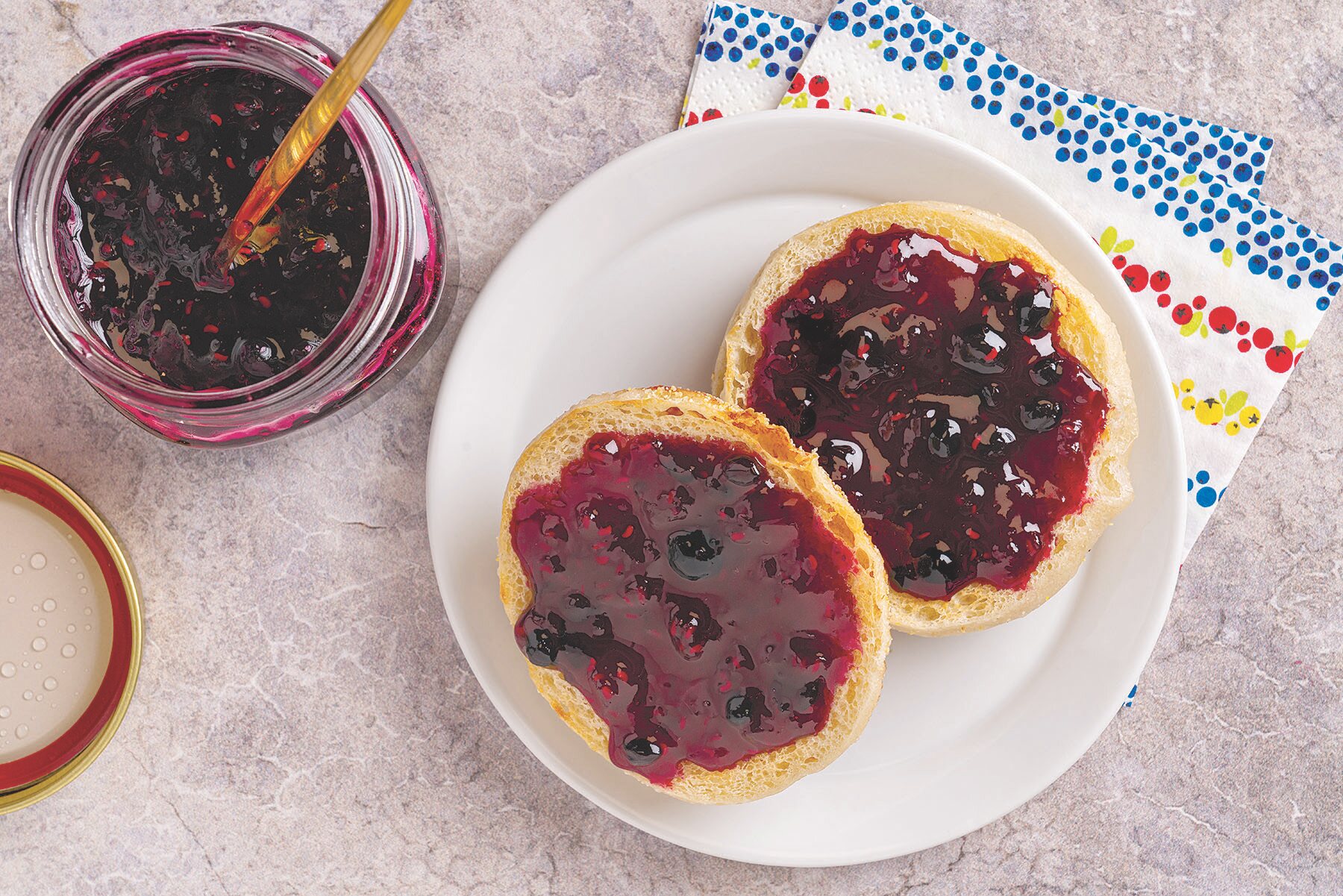 Two English muffin halves topped with wild blueberry and raspberry jam on a white plate, with a jar of jam and wooden spoon nearby