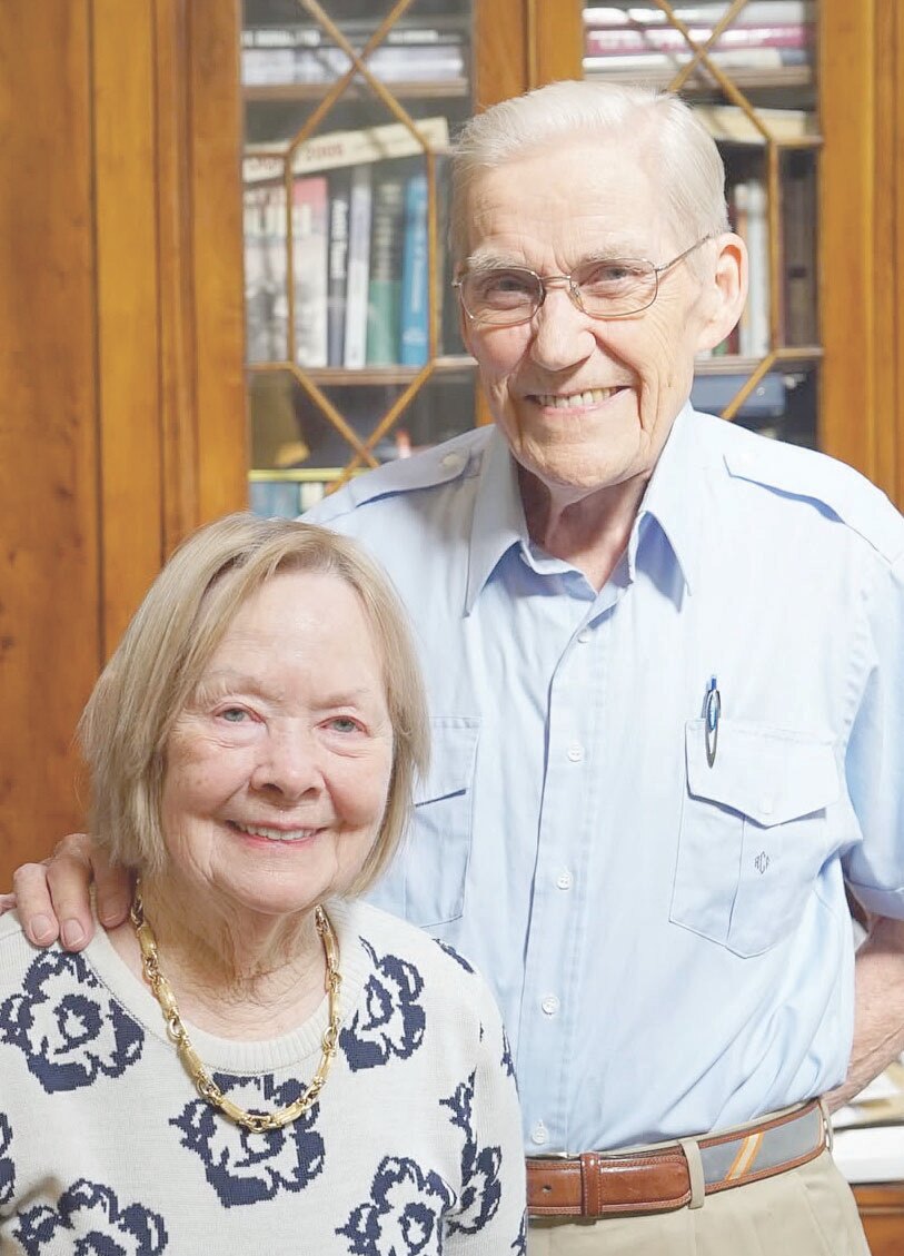A portrait of an elderly man and woman. The man is wearing a light blue shirt, glasses, and a brown belt, and the woman is wearing a white sweater with a blue floral pattern and a gold necklace. They are standing in front of a bookshelf.