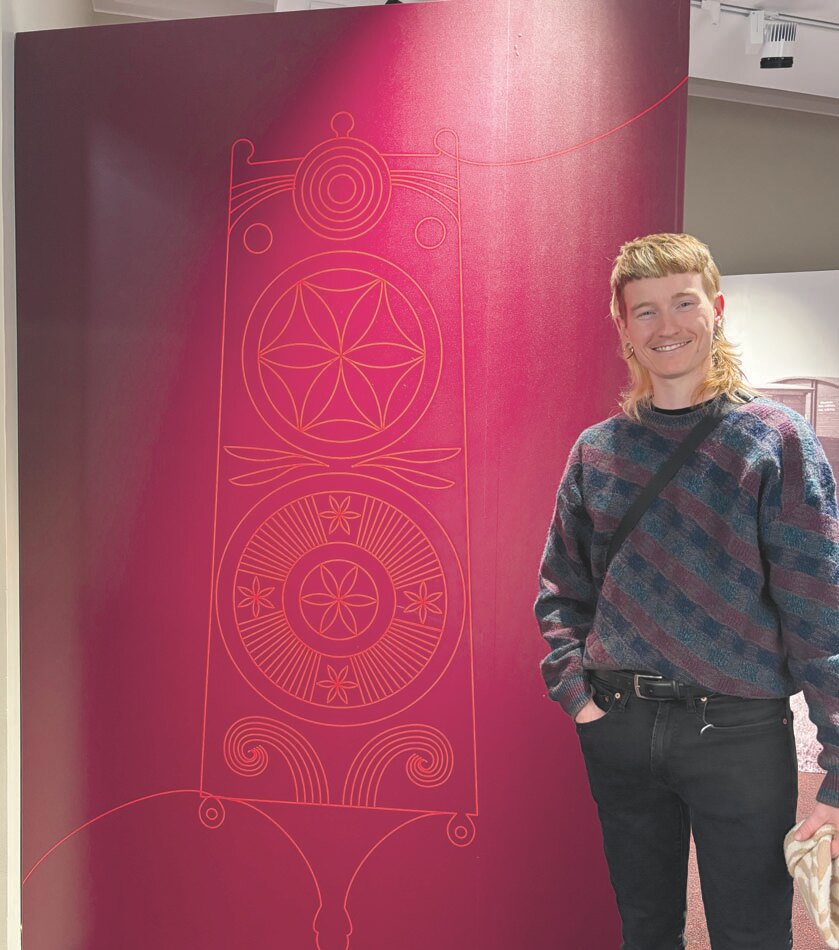 Author Gage Ker&auml;nen standing next to a tall red exhibit panel featuring traditional Karelian patterns