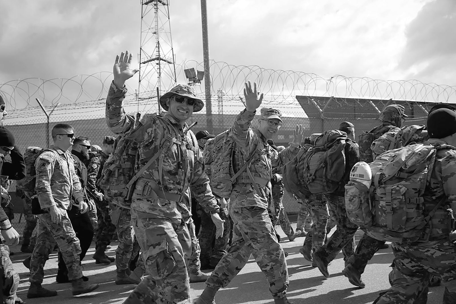 A group of soldiers marching with rucksacks; one soldier in the center front waves and wears sunglasses as they pass barbed wire and fencing.