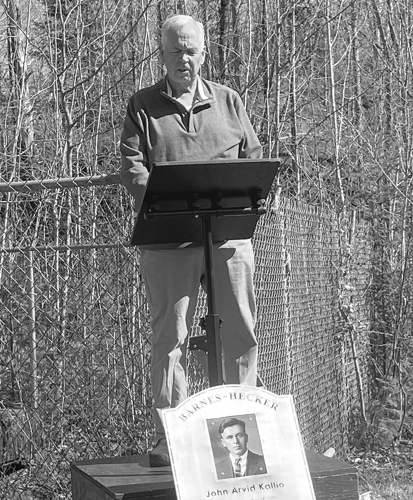 A black-and-white photo of a man speaking at an outdoor memorial podium with a Barnes-Hecker memorial plaque propped in front of it.