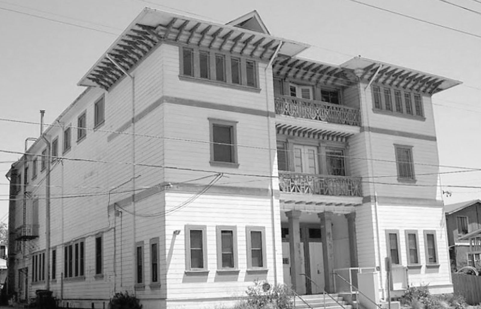 Black-and-white photograph of a three-story wooden building showing front and side facades, decorative wide eaves, a small central balcony/porch, multiple windows, utility lines crossing in front, and steps up to the main entrance.