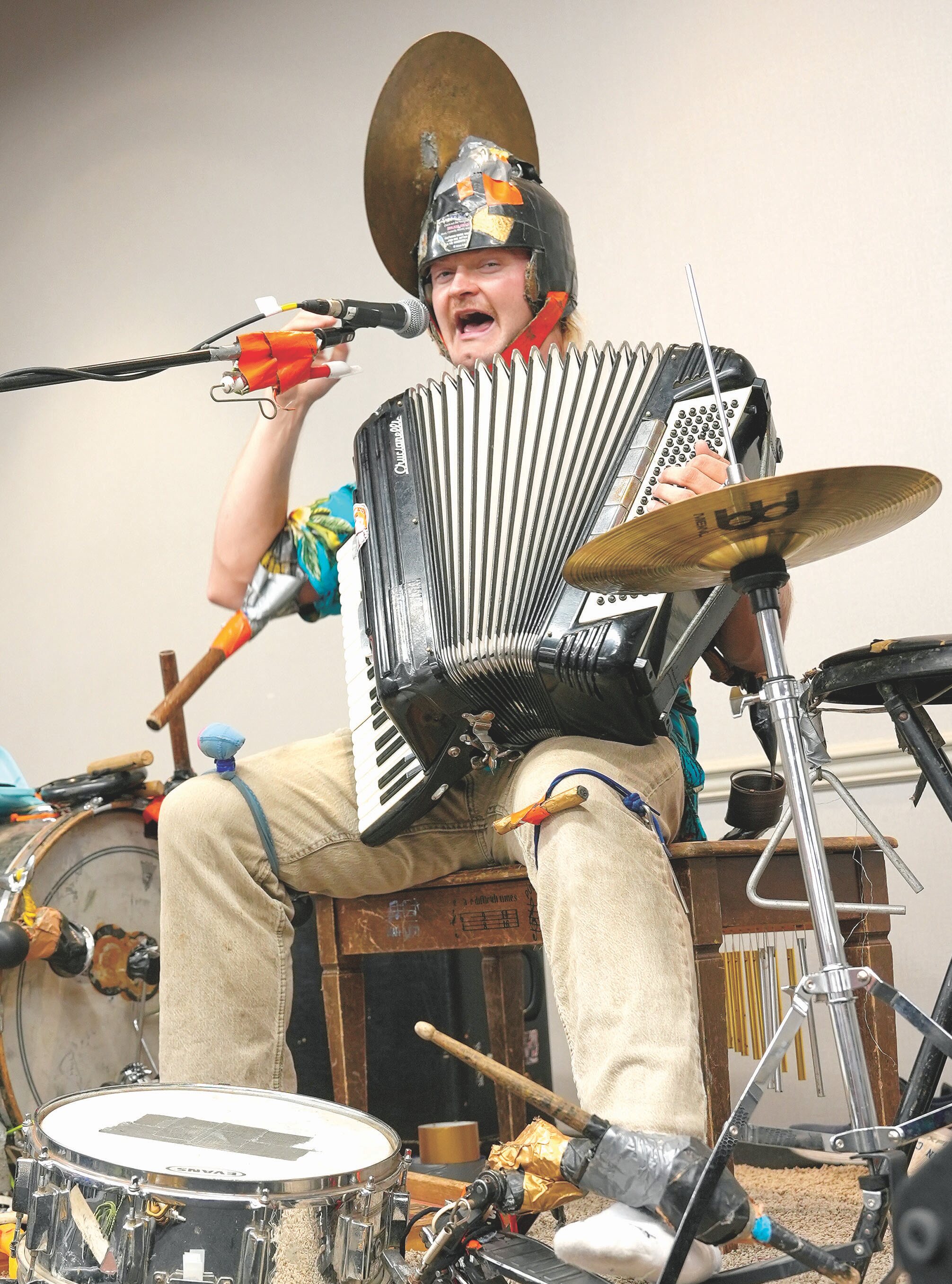 One-man-band performer playing an accordion and percussion, singing into a microphone.