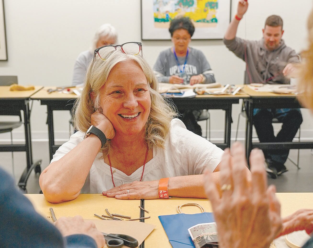 A smiling woman seated at a workshop table, leaning on her hand while others participate in the background.