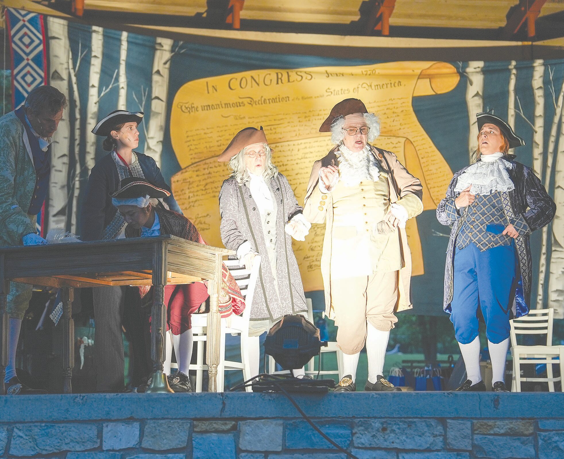 Stage performance with actors in colonial-era costumes in front of a backdrop reading 'IN CONGRESS'.