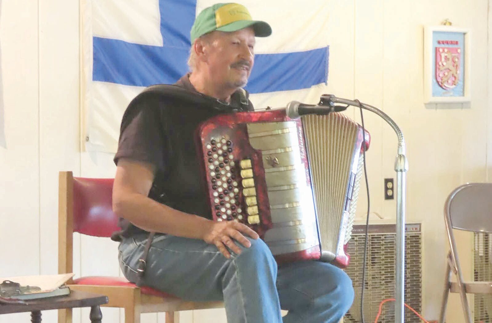 Man seated playing a red accordion with a Finnish flag in the background