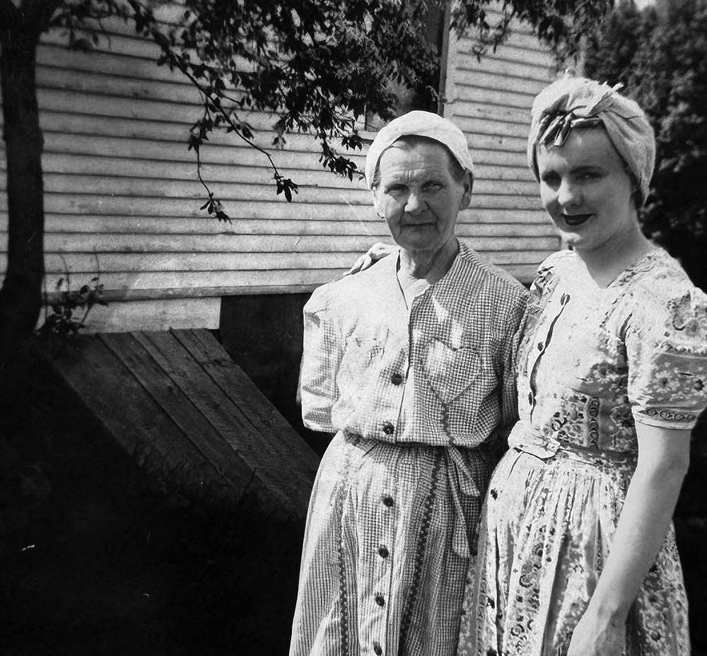 Black-and-white historic photo of two women standing side by side in front of a wooden building; an older woman on the left and a younger woman on the right wearing a headscarf.