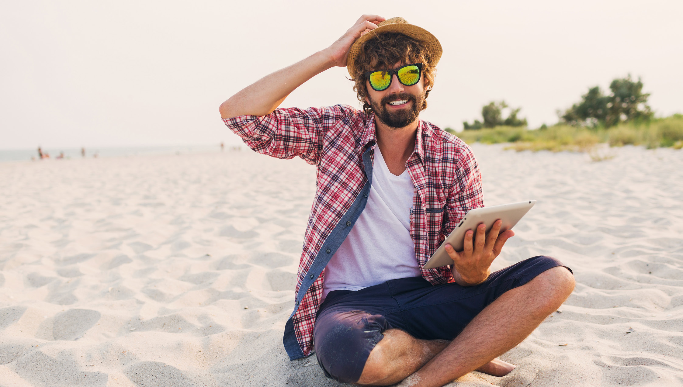 A bearded man in a straw hat and sunglasses sitting on a beach holding a tablet
