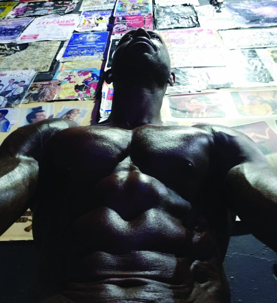 Low-angle photograph of a muscular, shirtless man looking upward against a wall of posters