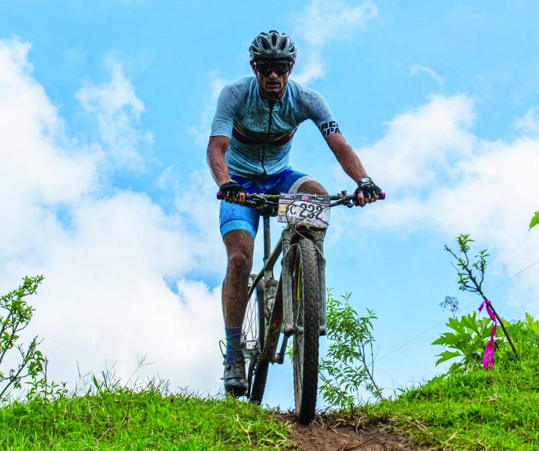 Cyclist riding a mountain bike downhill on a grassy slope under a blue sky with clouds.