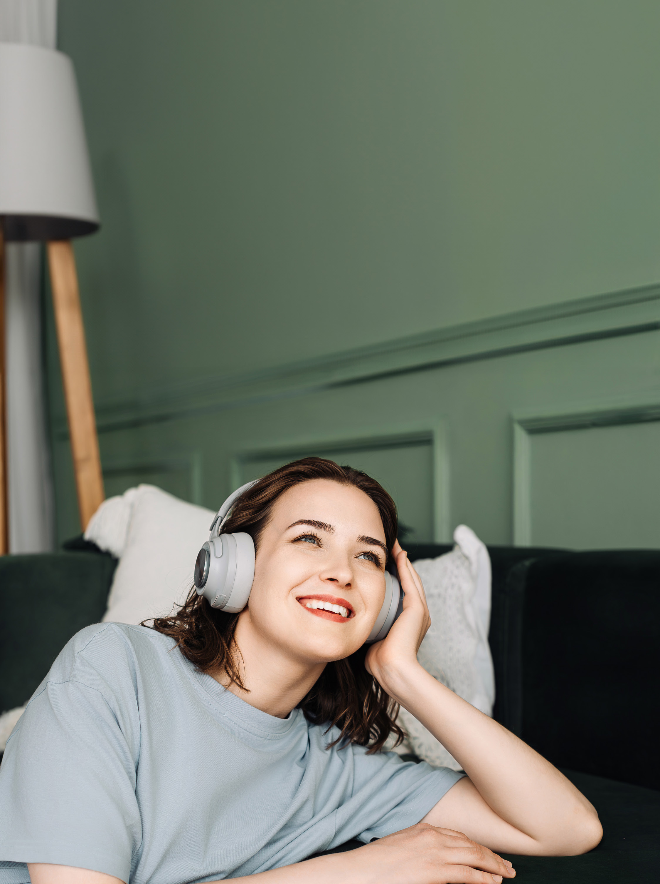 Photograph of a smiling woman reclining on a couch wearing over-ear headphones, looking up and holding one earcup; green paneled wall, a floor lamp and pillows visible behind her.