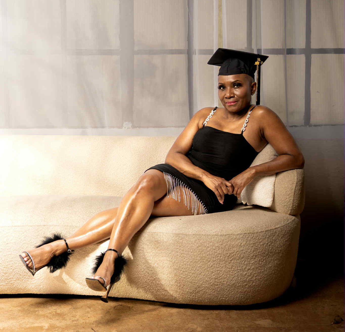 Woman seated on a curved beige sofa wearing a black dress and a graduation cap, smiling with one leg crossed.