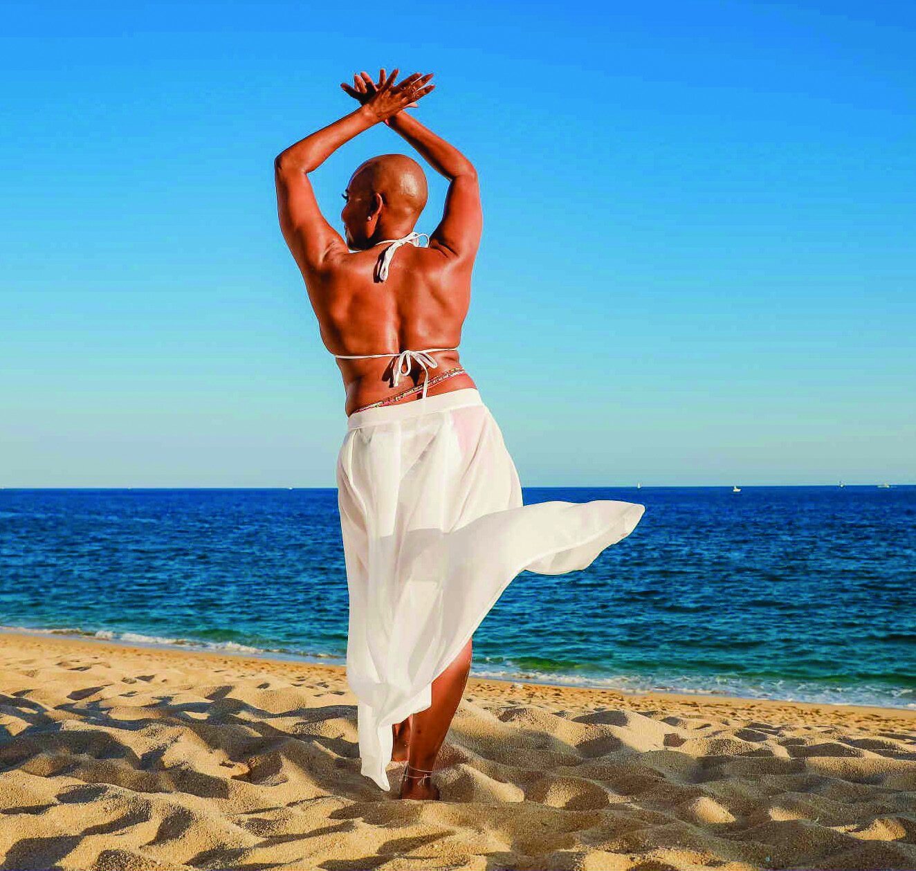 Person standing on a sandy beach facing the ocean with arms raised overhead, wearing a flowing white skirt that billows in the breeze.