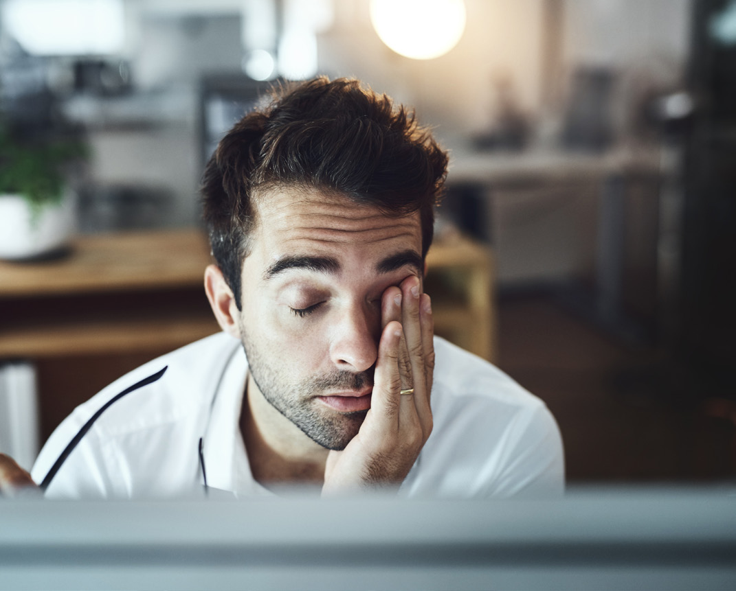 A photograph of a man looking tired, rubbing his eyes while working at a computer desk.