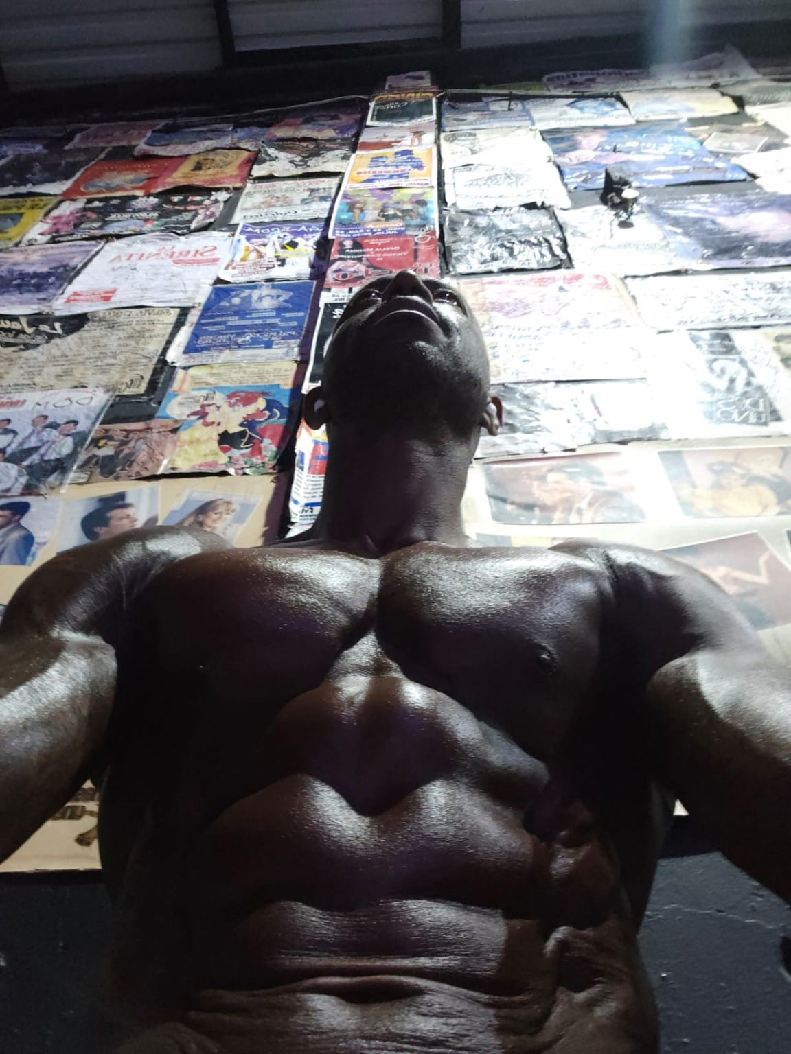Muscular man photographed from below, chest and abs prominent against a wall covered in posters