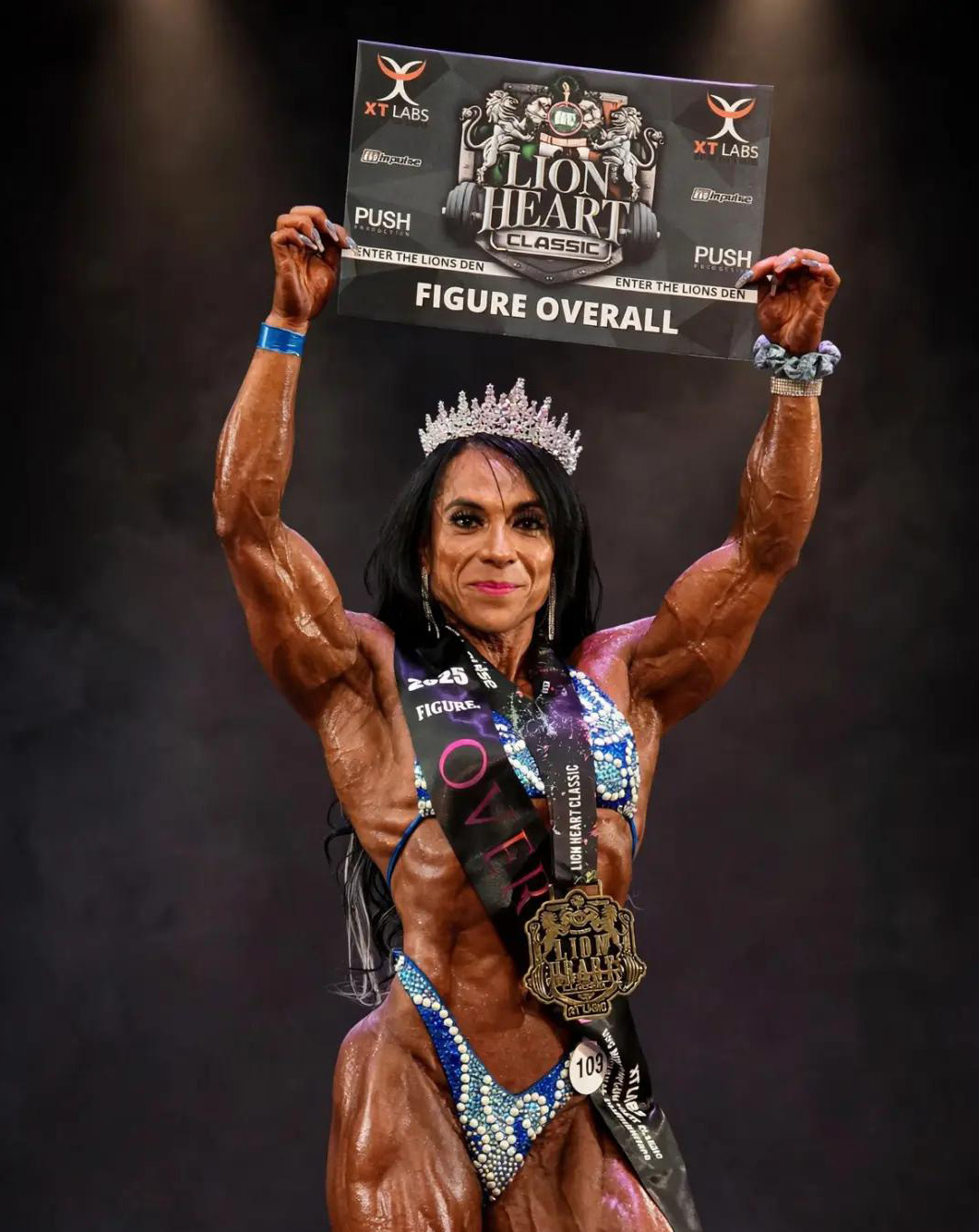 Maria Pe&ntilde;afiel on stage holding a 'FIGURE OVERALL' plaque above her head, wearing a crown and winner's medal