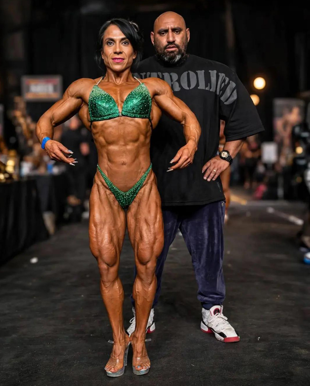Full-body photo of Maria Pe&ntilde;afiel standing on a competition floor in a green bikini, posing with her coach Gunther Xtreme standing behind her